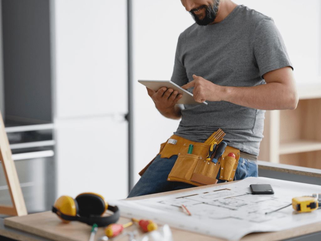 A smiling tradesperson in a grey t-shirt and tool belt using a digital tablet while leaning on a table with building plans, tools, and safety gear in a modern workspace.