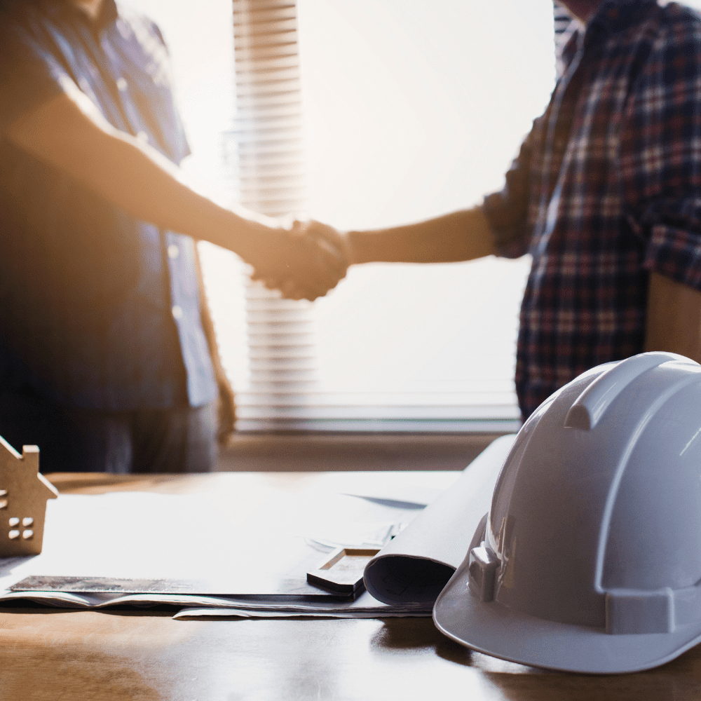 Builders shaking hands over house plans with safety helmet on desk